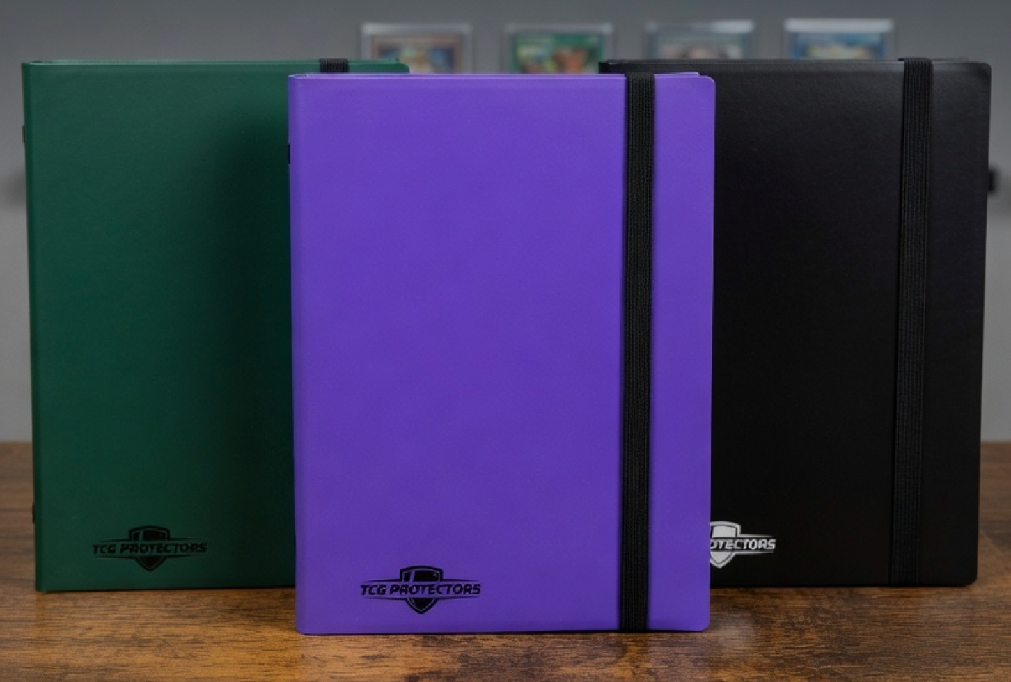 Three TCG Protectors binders in green, purple, and black on a wooden surface with a blurred background.